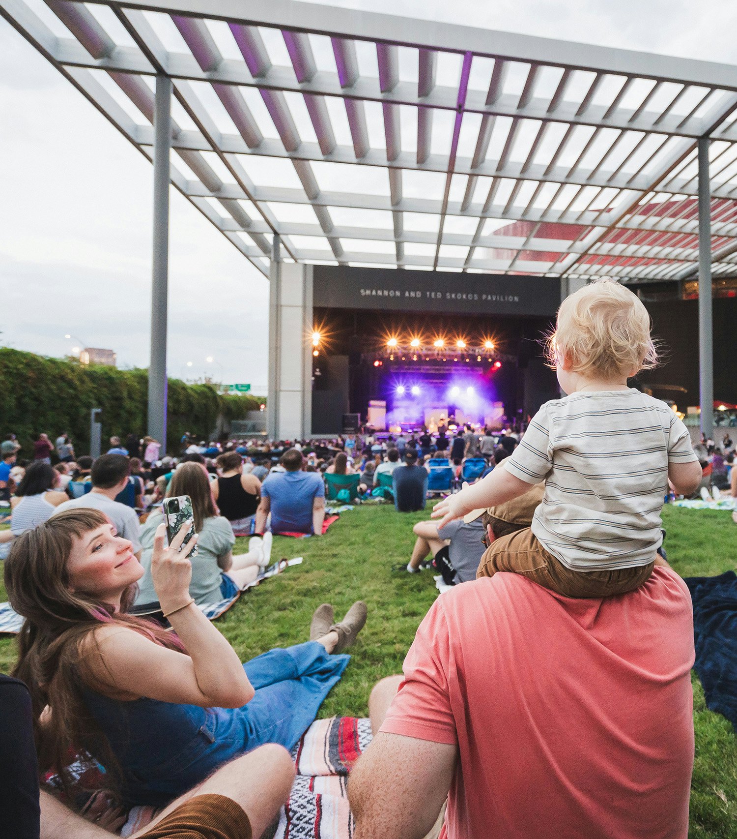 A man, woman and young child sitting on the grass in front of the stage at Annette Strauss Square in Dallas.